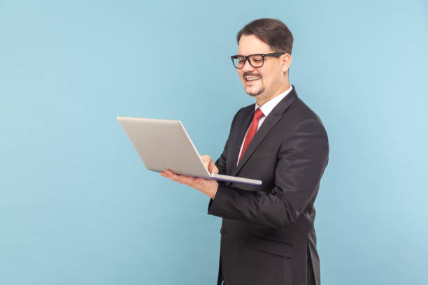 istock Smiling cheerful man standing and holding notebook, working on laptop online and joying his work. 1491350923