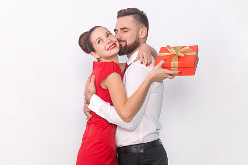 Portrait of romantic woman and man in elegant clothes hugging each other, female holding red present box, husband kissing his wife. Indoor studio shot isolated on gray background.