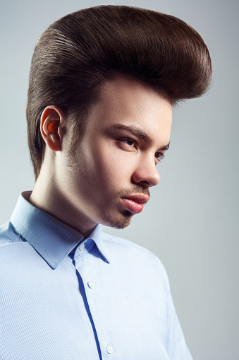 Side view portrait of handsome confident man with mustache and beard with retro classic elvis presley hairstyle, looking away, wearing blue shirt. Indoor studio shot isolated on gray background.