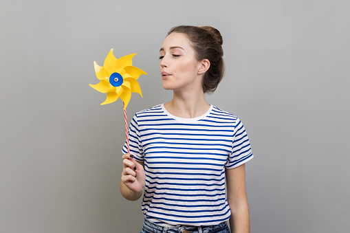 Origami hand mill. Portrait of carefree childish woman wearing striped T-shirt blowing at paper windmill, playing with pinwheel toy on stick. Indoor studio shot isolated on gray background.
