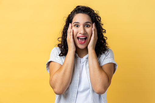 Portrait of surprised woman with dark wavy hair with mouth open in amazement, expressing shock, astonishment, keeping hands on his head. Indoor studio shot isolated on yellow background.