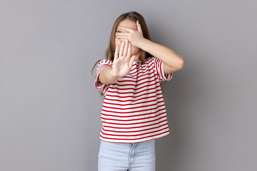 Portrait of sacred little girl wearing striped T-shirt closing eyes with palm of hand, stretching out another hand forward, showing stop gesture. Indoor studio shot isolated on gray background.