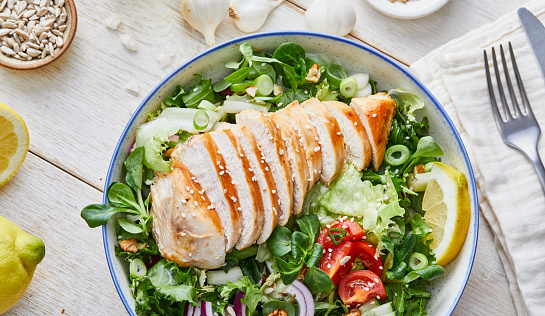 Delicious homemade gourmet healthy chicken salad bowl, with Spanish onion, cherry tomato, lemon and olive oil, on a garlic and lettuce green salad, served in a rustic handmade ceramic bowl, with seasoning and herbs on a white rustic natural wooden kitchen or restaurant table, representing a healthy lifestyle, wellbeing and body care, top view with a copy space