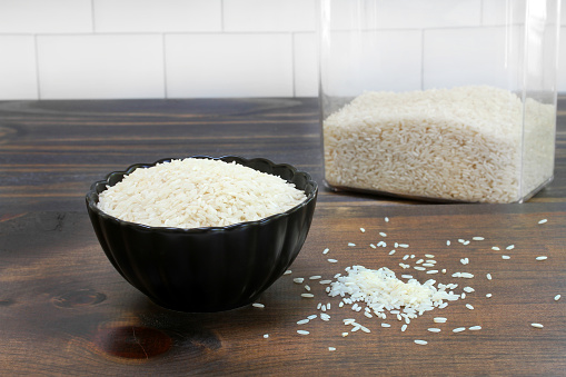 Raw long grain white rice in a black bowl and on a wooden table.