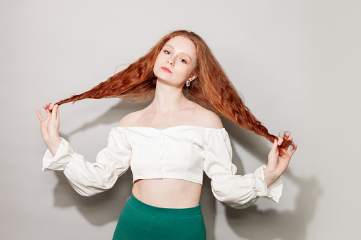 Studio portrait of a young attractive cheerful red-haired woman in a white blouse against a gray background