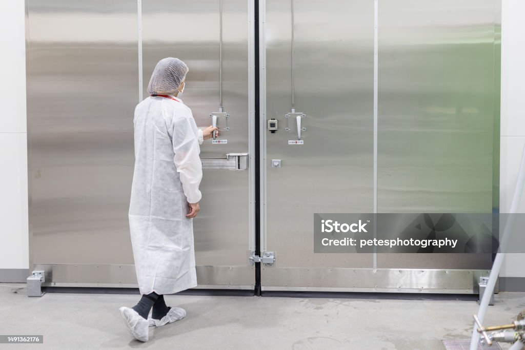 Female in white clean suit opening a large aluminum door A rear view of a female worker in white clean covers opening a large industrial door. Refrigerator Stock Photo