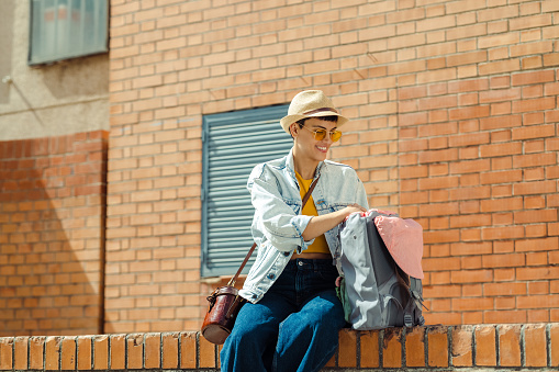 Modern short hair woman sitting on the brick wall and checking backpack