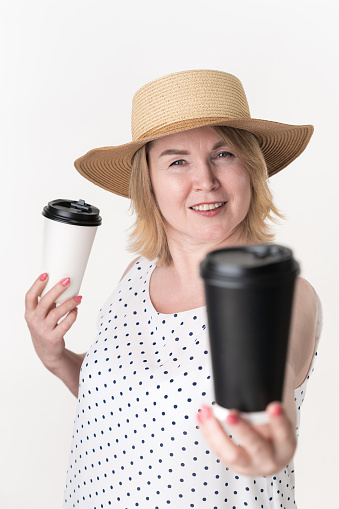 Playful blonde woman holding black and white disposable paper cup for coffee in different hands. Choice concept tea or coffee. Middle aged adult female wearing in white dress and straw hat