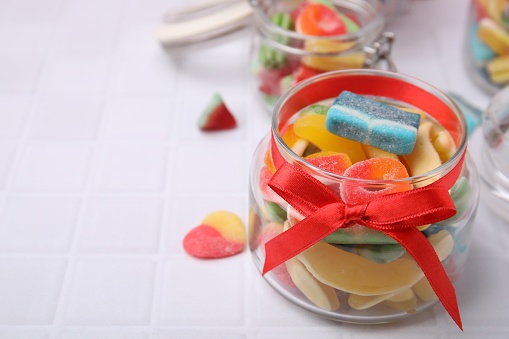 Glass jars with tasty colorful jelly candies on white tiled table. Space for text
