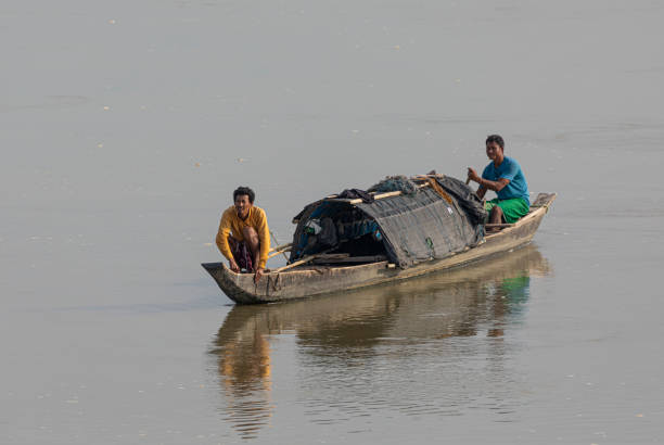 2 April 2023 - Assam, India. Two fishermen in a small boat in the Brahmaputra River, Assam, India. There is fishing net draped across the roof of the boat, and bamboo poles for testing the depth of the water.