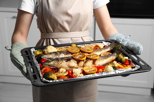 Woman holding baking tray with sea bass fish and vegetables in kitchen, closeup