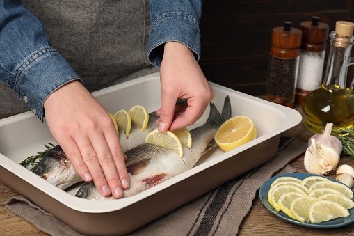 Woman putting pieces of lemon in raw sea bass fish at wooden table, closeup
