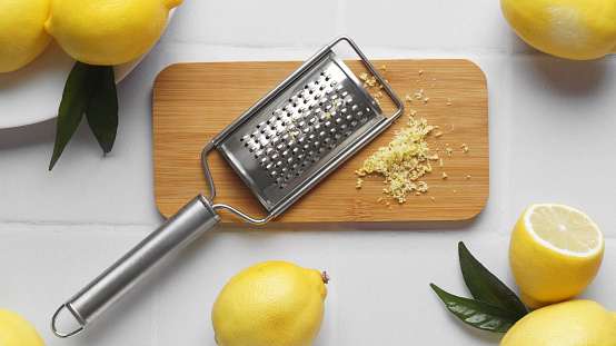Wooden board with grater and fresh lemon zest on white tiled table, flat lay