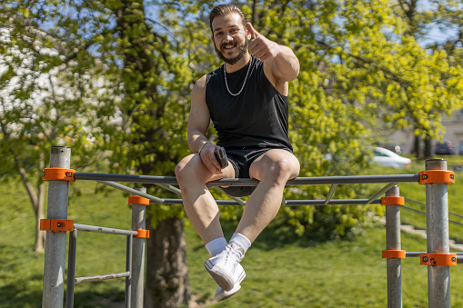 Athletic man in sportswear raises thumbs up gives positive feedback likes good. Guy on playground sits on horizontal bar. Sports health, fitness routine, workout. Strength and motivation. Outdoor gym