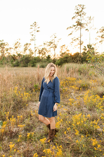A Happy & Joy-Filled 30-Year-Old American Woman with Short Straight Blond Hair & Baby Blue Eyes Wearing a Blue Denim Dress & Brown Leather Cowboy Boots Enjoy Time Outdoors in a Field of Yellow Wildflowers & Pampas Grass at Golden Hour in Jupiter Farms, Florida.