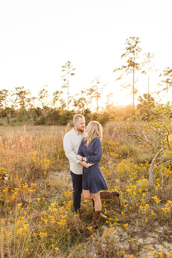 A Happy Young American Couple with Short Blond Hair & Baby Blue Eyes Wearing Denim & Brown Leather Cowboy Boots, Snuggling & Enjoying Time Together Outdoors in a Field of Yellow Wildflowers & Pampas Grass at Golden Hour in Jupiter Farms, Florida.