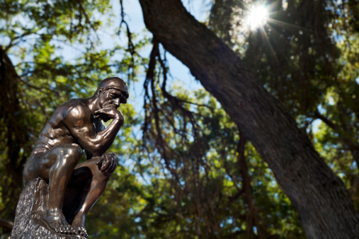 View of a small bronze replica of Rodin's The Thinker statue, sitting in a spot of light in a forest.  The Thinker is often used to represent philiosphy.
