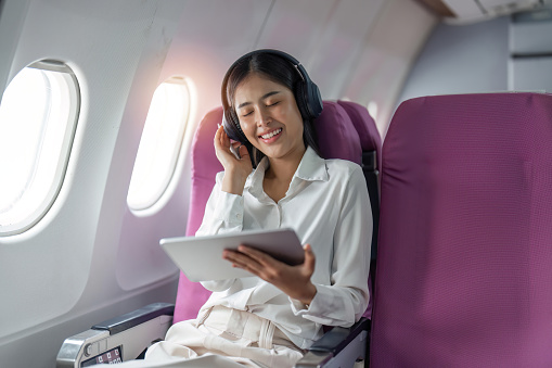 Young asian woman in wireless headphones listening to music and smiling during fly in airplane, travel concept.