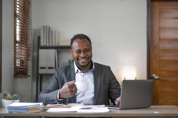 istock Young African black mansmiling businessman drinking coffee and using laptop in office, sitting at desk 1491403601