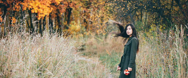 Female beauty portrait surrounded by vivid foliage. Dreamy beautiful girl with long natural black hair in full growth on background with colorful leaves. Fallen leaves in girl hands in autumn forest.