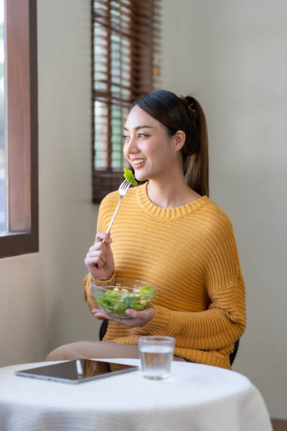 istock Happy young Asian woman eating salad at home and using laptop computer. 1491418410