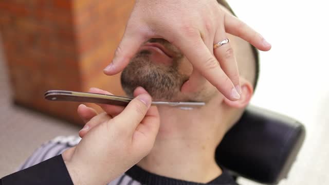 Barber Man is shaving client with a vintage straight razor in a barbershop. Classic shave by stainless steel straight edge razor