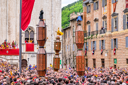 Gubbio, Umbria, Italy, May 15 -- The start of the race during the ancient Festa dei Ceri in the historic heart of the medieval town of Gubbio in Umbria. The celebration consists of a race of three wooden columns weighing almost 300 kg each, called Ceri, carried on the shoulders of dozens of bearers, on top of which are placed the statues of Sant'Ubaldo (Saint Ubaldo) protector of Gubbio, San Giorgio (Saint George) and Sant'Antonio Abate (Saint Anthony). The race develops along the streets and alleys of Gubbio up to the summit of Monte Ingino where the Basilica of Sant'Ubaldo is located. This religious celebration, much loved by the whole community of Gubbio, is considered one of the oldest in Italy and in the world and its origins date back to the 12th century. In the photo: The three wooden columns of the Ceri with Sant'Ubaldo, San Giorgio and Sant'Antonio start the race from the Piazza Grande in the heart of Gubbio. Image in high definition quality.
