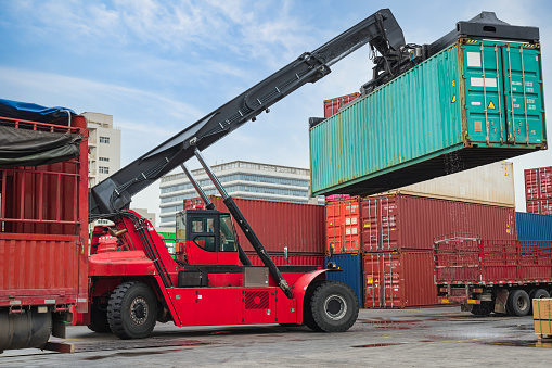 Transferring freight containers from train to truck at a city intermodal hub. A modern container stacker loads a freight container onto a truck on a hardstand container terminal. In the background are rail wagons (freight cars) loaded with a variety of containers. Logos and trademarks have been removed.
