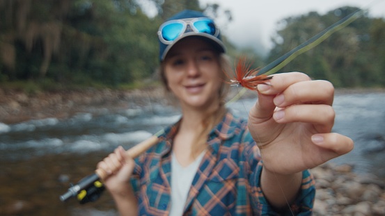 Woman angler on the river. Young woman holds the fishing fly in her hand and shows the lure to camera