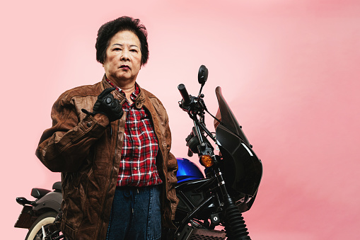 Senior woman wearing leathers standing with motorcycle, posing in studio