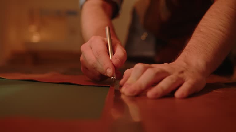 Craftsman shoemaker cutting genuine leather with knife scalpel on table. Leatherworker at work making handmade shoes.