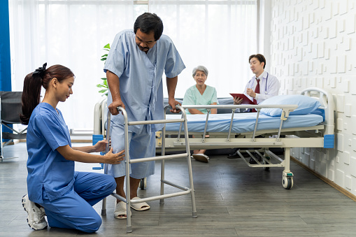 Young Female surgeon checking patient's knee condition after surgery in hospital room, Help Patient male training to walk with walker equipment