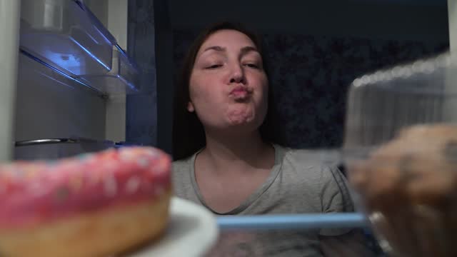 Adult woman opens fridge and grabs doughnut eating at night