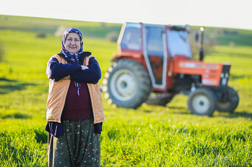 senior farmer woman with his arms tied standing in front of his orange tractor