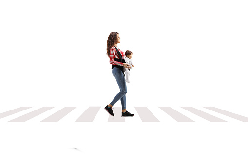 Full lenght shot of a young mother with a babay in a carrier walking at pedestrian crossing isolated on white background