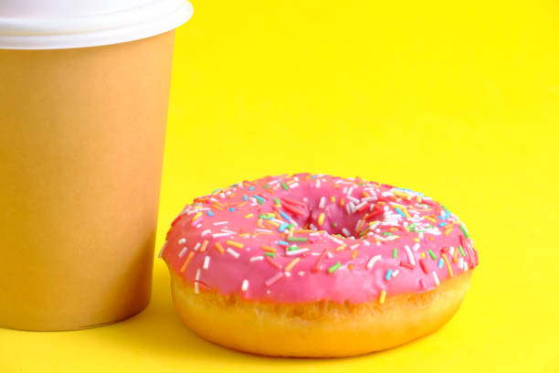 istock Disposable coffee cup and doughnut with pink icing yellow background. 1491461104
