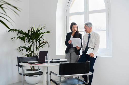 Two business people meeting near a window looking at a document. There is paperwork and a laptop on the table, formal business wear.