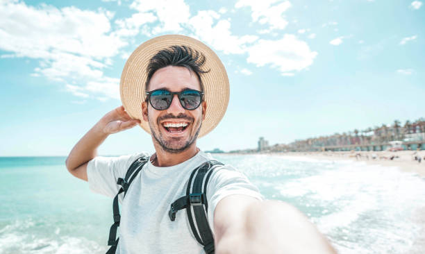 uomo felice con cappello e occhiali da sole che scattano foto selfie con smartphone in spiaggia - viaggiatore allegro che si diverte fuori - bel ragazzo che sorride alla macchina fotografica - australia photographing camera beach foto e immagini stock