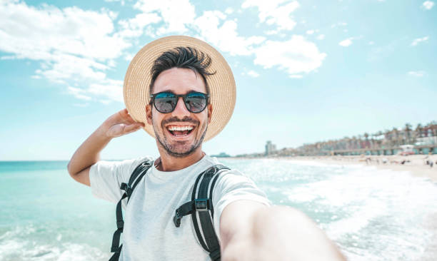 ビーチでスマートフォンで自分撮り写真を撮る帽子とサングラスを持つ幸せな男性 – 外で楽しむ陽気な旅行者 – カメラに微笑むハンサムな男 - australia photographing camera beach ストックフォトと画像