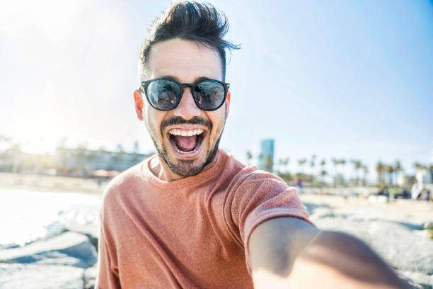外のカメラに微笑む幸せな若い男 – スマート携帯電話デバイスで自分撮りをするハンサムな男 – 観光、技術、人々のコンセプト - australia photographing camera beach ストックフォトと画像
