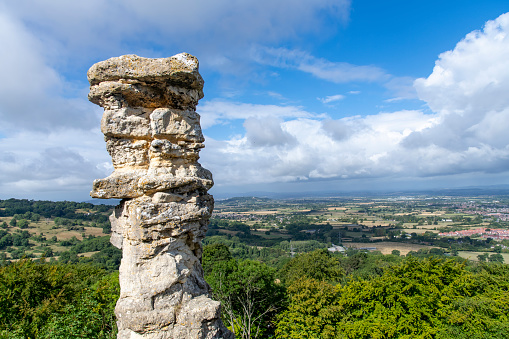Close up view of limestone rock formation Devils Chimney against backdrop of panoramic view from Leckhampton Hill, Cheltenham, UK along the public footpath Cotswold Way overlooking the Cotswolds