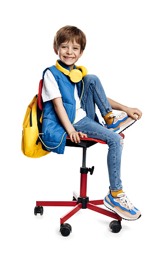 Happy little boy wearing headphone and yellow backpack sitting on red chair on white background. Back to School