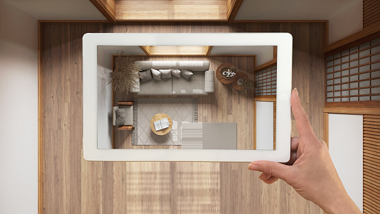 Augmented reality concept. Hand holding tablet with AR application used to simulate furniture and design products in empty interior with parquet floor, japandi living room, top view