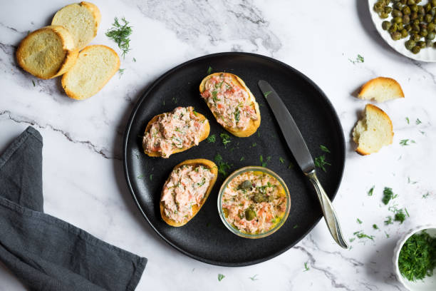 Salmon rillettes, mousse, pate and toasts on wooden background Salmon rillettes, mousse, pate and toasts on wooden background. Bowl of salmon pate served with butter, sliced bread, capers, vintage knife and herbs. Top view. High quality photo. tuna pate stock pictures, royalty-free photos & images