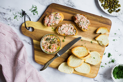 Salmon rillettes, mousse, pate and toasts on wooden background. Bowl of salmon pate served with butter, sliced bread, capers, vintage knife and herbs. Top view. High quality photo.