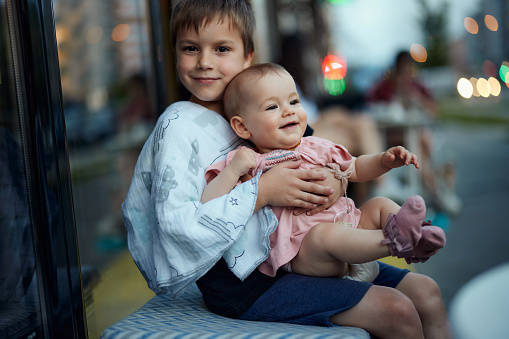 Smiling older brother holding his baby sister in lap outdoors.