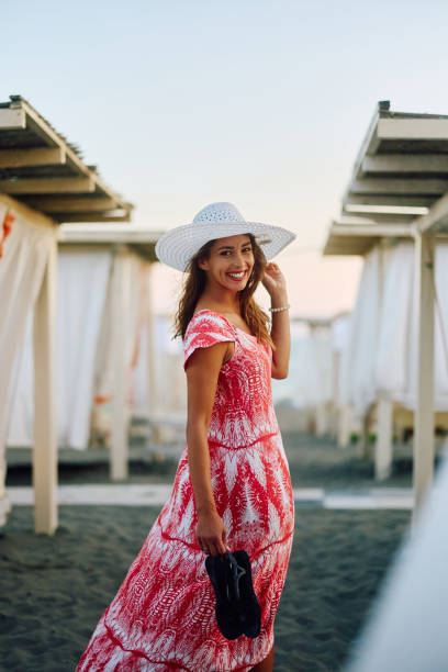 Happy woman wearing colorful dress and sun hat while walking on the beach on summer day and looking at camera.