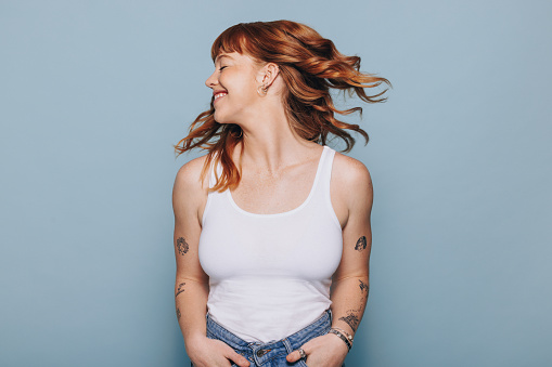 Happy woman flipping her ginger hair while standing against a blue background. Young woman having fun in casual clothing.