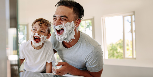 Father and son having fun in a bathroom, laughing happily with shaving foam on their faces. Young single dad taking a moment to bond and share moments of joy with his boy on father's day.
