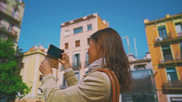 Slow motion of smiling female tourist photographing in city on sunny day. Woman is using smart phone at town square. She is exploring during vacation.